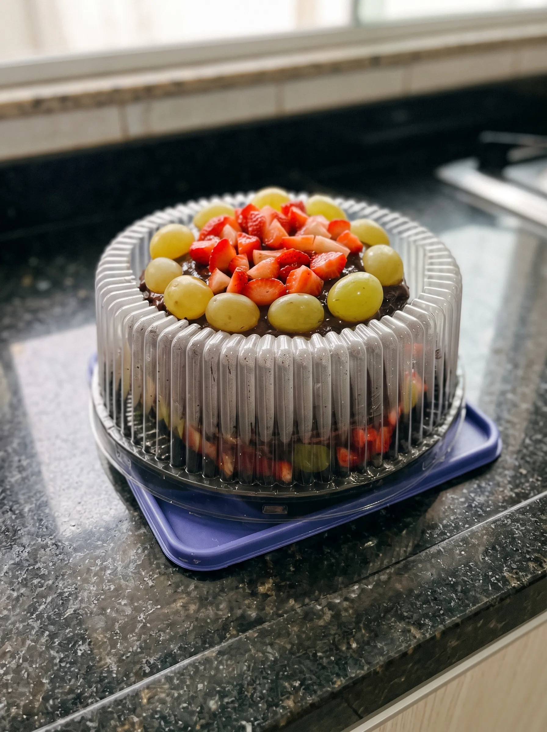 Bolo de chocolate coberto de morangos e uvas, numa embalagem transparente sobre a bancada